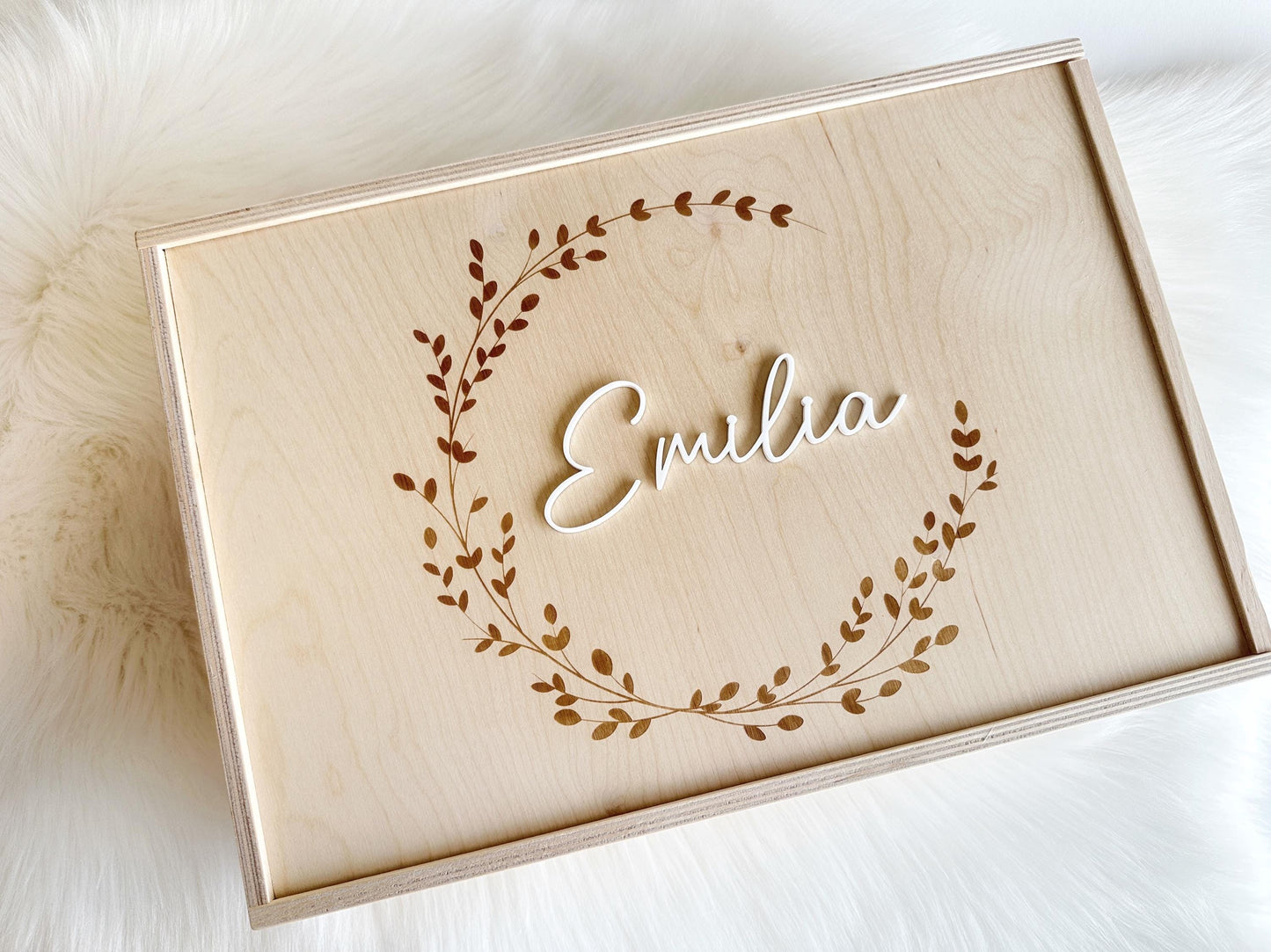 Mittig platziert: personalisierte Holzkiste mit Schiebedeckel, Gravur und Namen aus Acrylglas. Die Kranzgravur ist in runder Form und der Name ist mittig platziert. Die Erinnerungsbox steht auf einem weißen Fell.