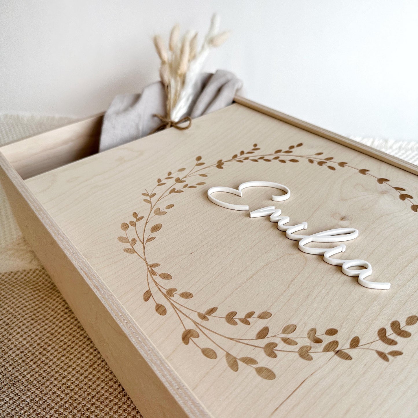Schräge Aufnahme der der Erinnerungsbox für Babys. Der Schiebedeckel istleicht geöffnet und mit einem schmalen Trockenblumengesteck gefüllt. Die saubere und detailierte Gravur vom Kranz ist gut sichtbar.