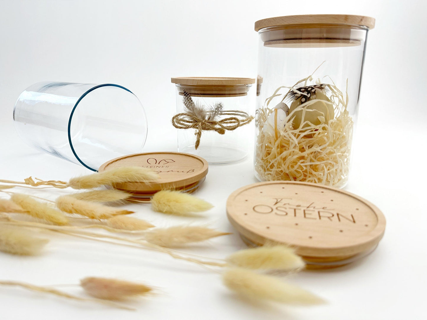 Seitliche Nahaufnahme der Vorratsgläser, im Vordergrund liegt ein gravierter Holzdeckel mit Ostermotiv, zwei Glaser stehen beispielhaft dekoriert dahinter, ein leeres Glas liegt seitlich links daneben, davor sind Trockenblumen dekoriert, passend zu Ostern und zum Frühling
