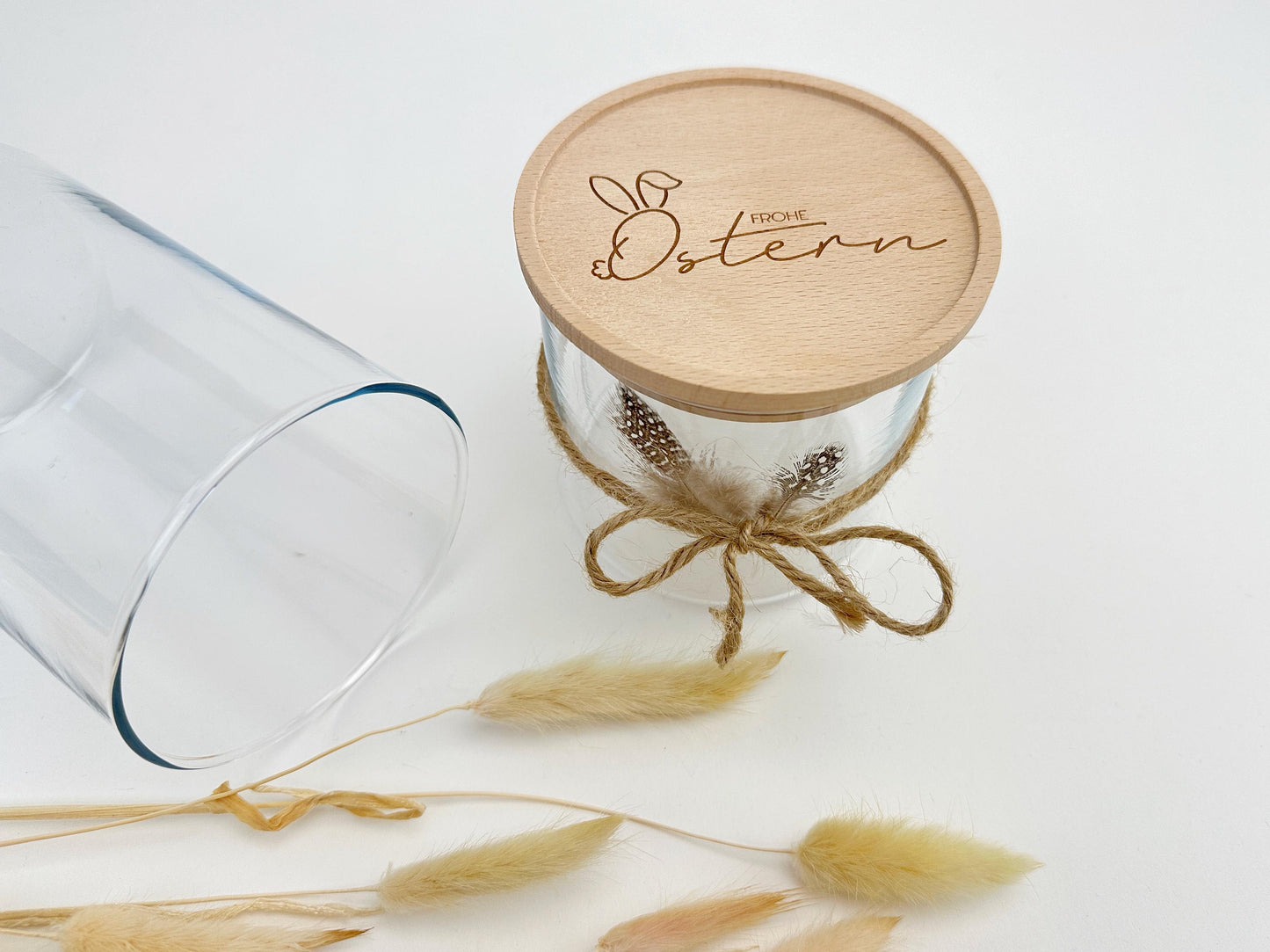 Oster-Vorratsglas mit graviertem Holzdeckel 'Frohe Ostern'. Dekoriert mit Juteband, Federn und Trockenblumen für eine natürliche, rustikale Osterdeko. Perfekt als nachhaltige Geschenkverpackung für Ostern, zum Befüllen mit Süßigkeiten oder kleinen Geschenken