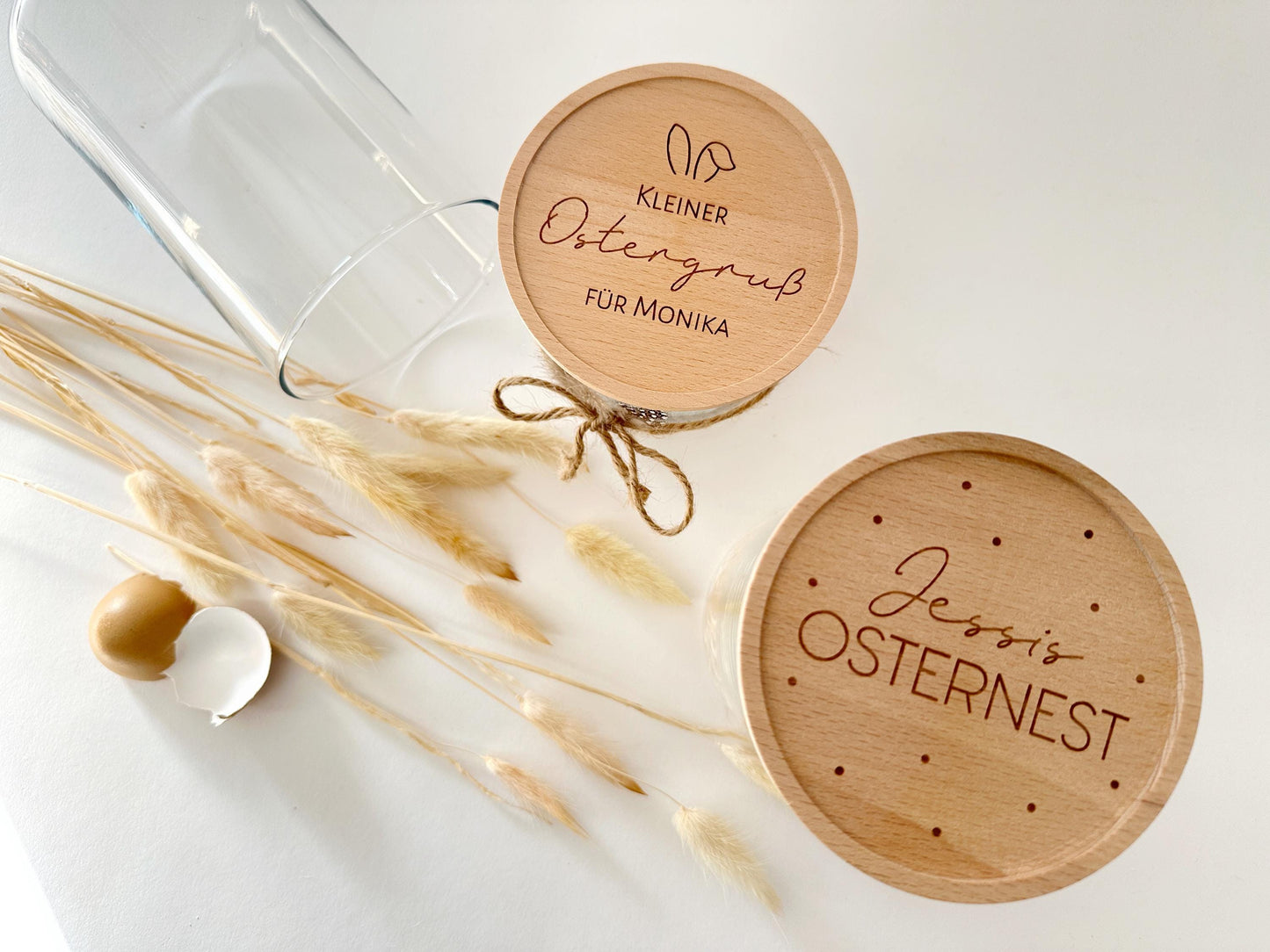 Das Bild zeigt zwei edle Vorratsgläser mit gravierten Holzdeckeln in einer stilvollen Osterdekoration. Eines der Gläser ist mit natürlichem Ostergras, einem Ei und Federn befüllt, während das andere mit einer rustikalen Juteschleife verziert ist. Die feinen Gravuren auf den Deckeln zeigen ein niedliches Osterhasenmotiv und elegante Schriftzüge. Drumherum sind kleine Deko-Eier und eine zerbrochene Eierschale arrangiert, was das frühlingshafte Osterambiente perfekt unterstreicht. 🌿🥚🐣