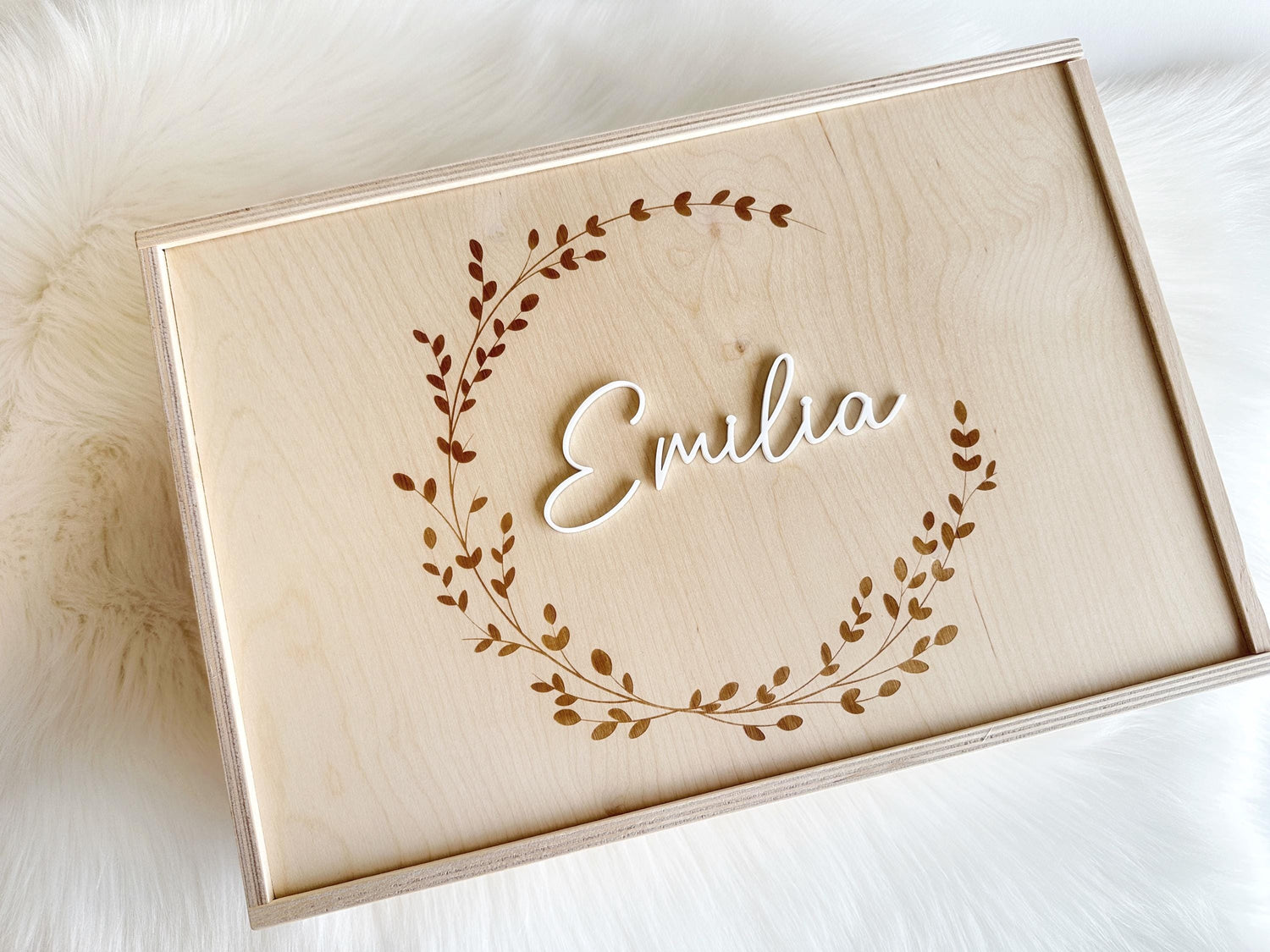Mittig platziert: personalisierte Holzkiste mit Schiebedeckel, Gravur und Namen aus Acrylglas. Die Kranzgravur ist in runder Form und der Name ist mittig platziert. Die Erinnerungsbox steht auf einem weißen Fell.