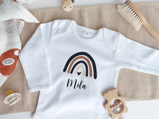 Mittig liegt ein weißer Baby langarm Body mit einem Regenbogen als Motiv. In der Mitte des kleinsten Bogens ist ein kleines Herz und darunter Platz für den Name. Der Body liegt auf einer hellbraunen Stoffbahn. Drumherum liegen Baby-Accessoires.