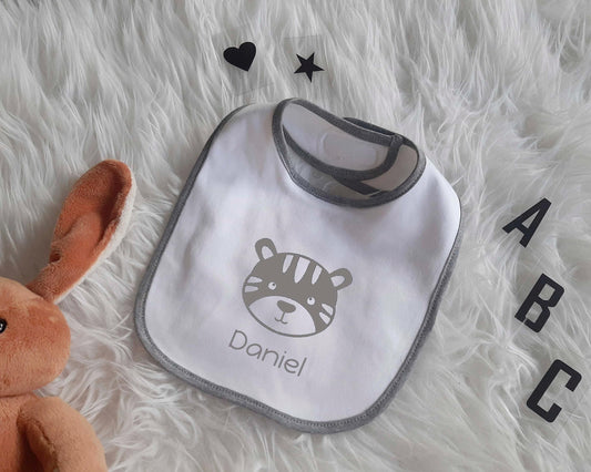 Baby Lätzchen mit Tiermotiv und personalisiertem Name.Der Sabberlatz liegt auf einem weißen Fell. Drumherum liegen Buchstaben und ein Herz und ein Stern. Unten links im Eck ist ein Stoffhase zu sehen. Ideales Geschenk zur Taufe oder Geburt.